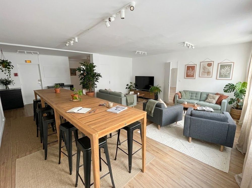 Salle de pause : grande table haute en bois avec tabourets, coin salon avec canapés et télévision